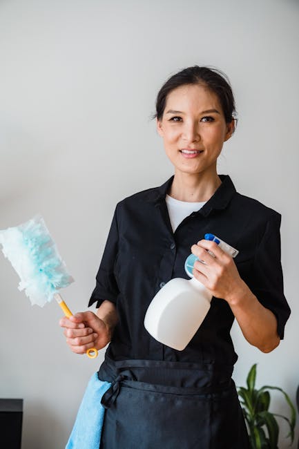 A professional cleaner dressed in a white protective suit, gloves, and a face mask is using a mop to clean large glass windows inside a modern building. The cleaning process involves surface cleaning
